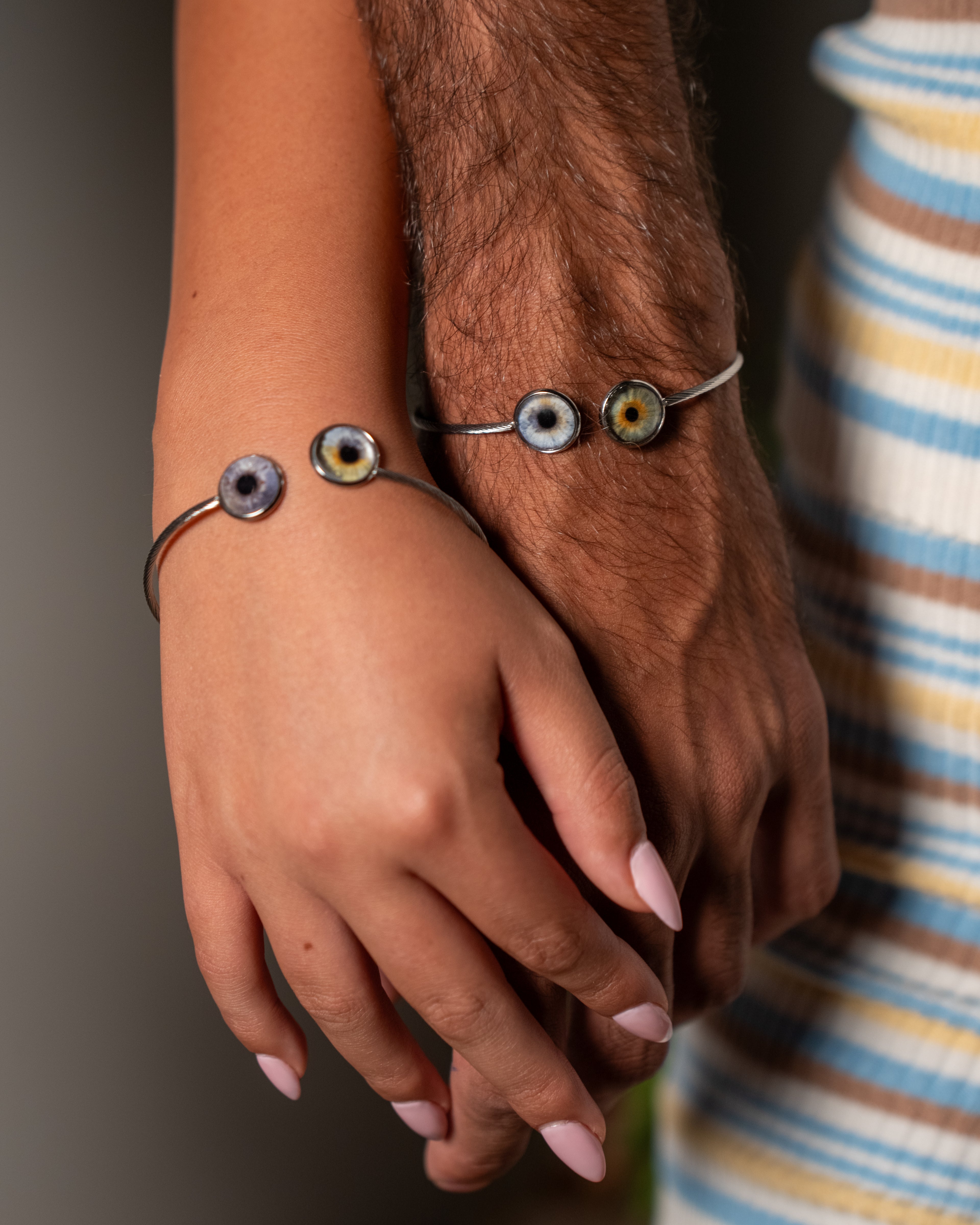 Two hands holding each other wearing matching bracelets with eye-shaped beads.